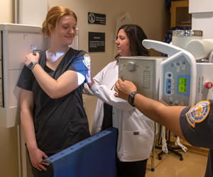 Radiology student preparing to use x-ray machine on another smiling radiography student with the instructor assisting